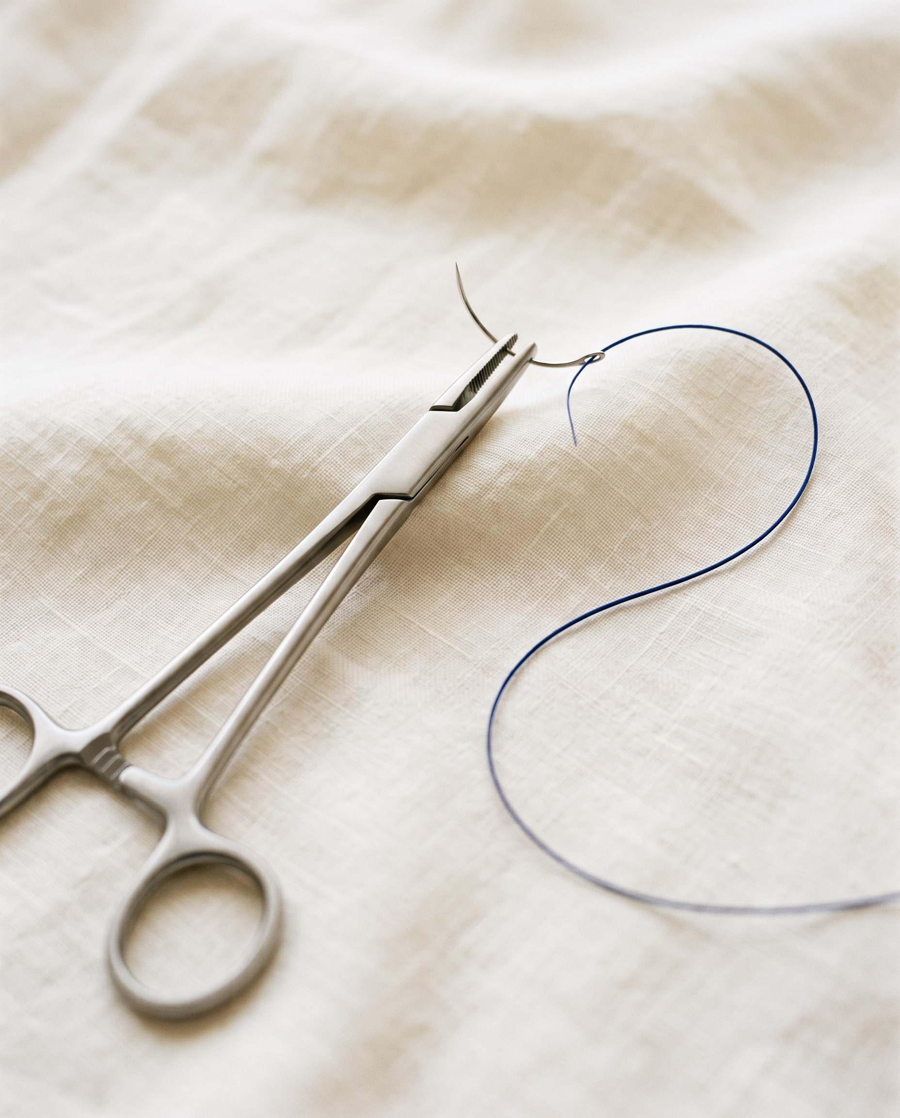 A needle holder clamping a curved suture needle threaded with fine blue monofilament, resting on ivory linen.